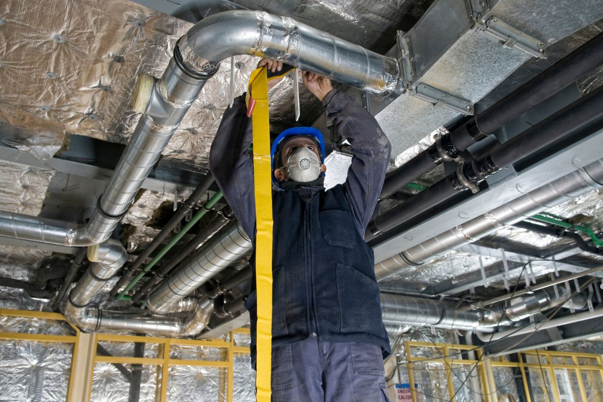  A man in safety gear and a mask holds a measuring tape, inspecting for mold in an HVAC system. 